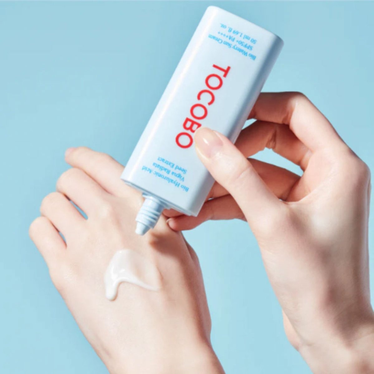 Hand applying cream from a Tocobo bottle against a light blue background